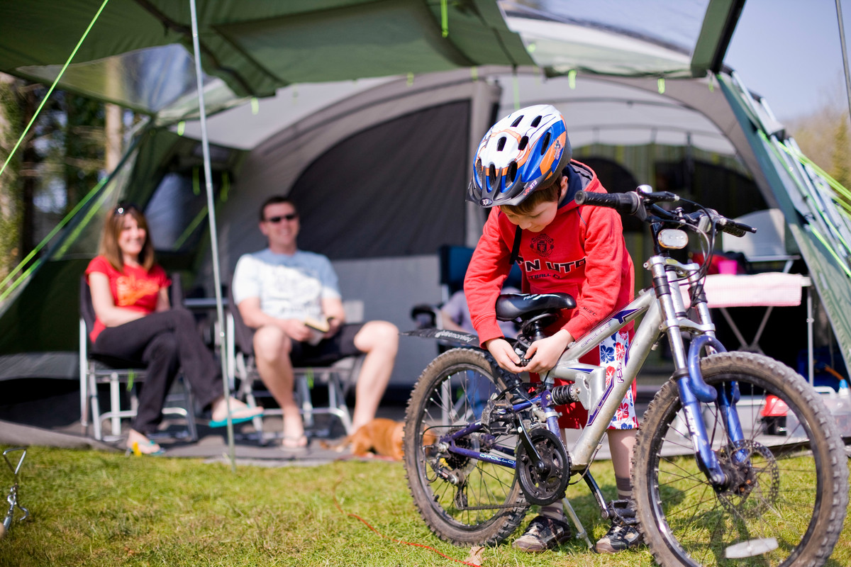 Boy with bicycle helmet leaning over bicycle adjusting lock as man and woman on deck chairs look on smiling.